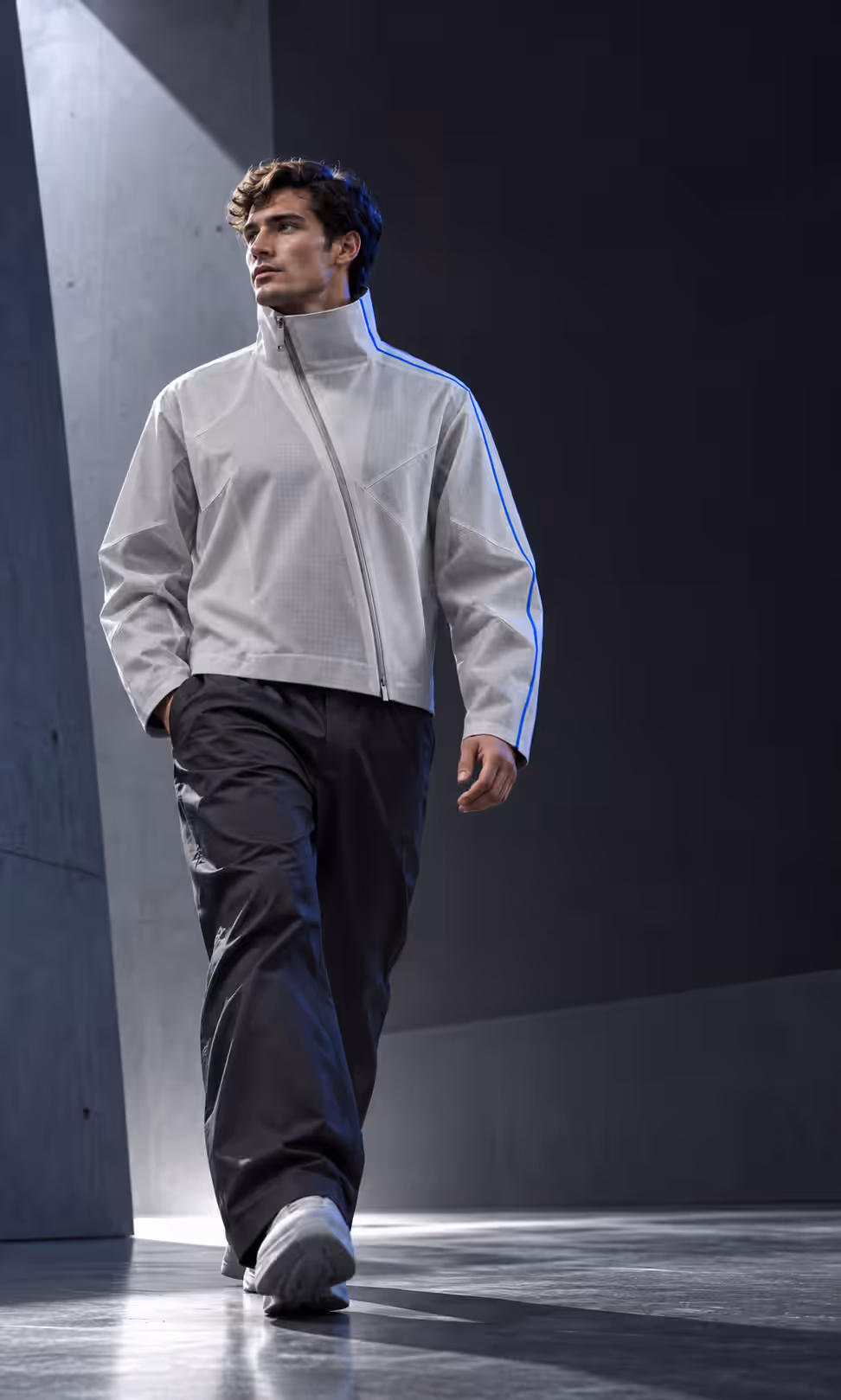 A man in the light grey jacket and dark trousers walking through a concrete corridor